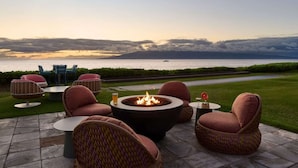 An outdoor seating area with a fire pit, wicker chairs, and a view of the sea at sunset. - Bungalow with Garden View + Kitchen + Pool Access (Lahaina)