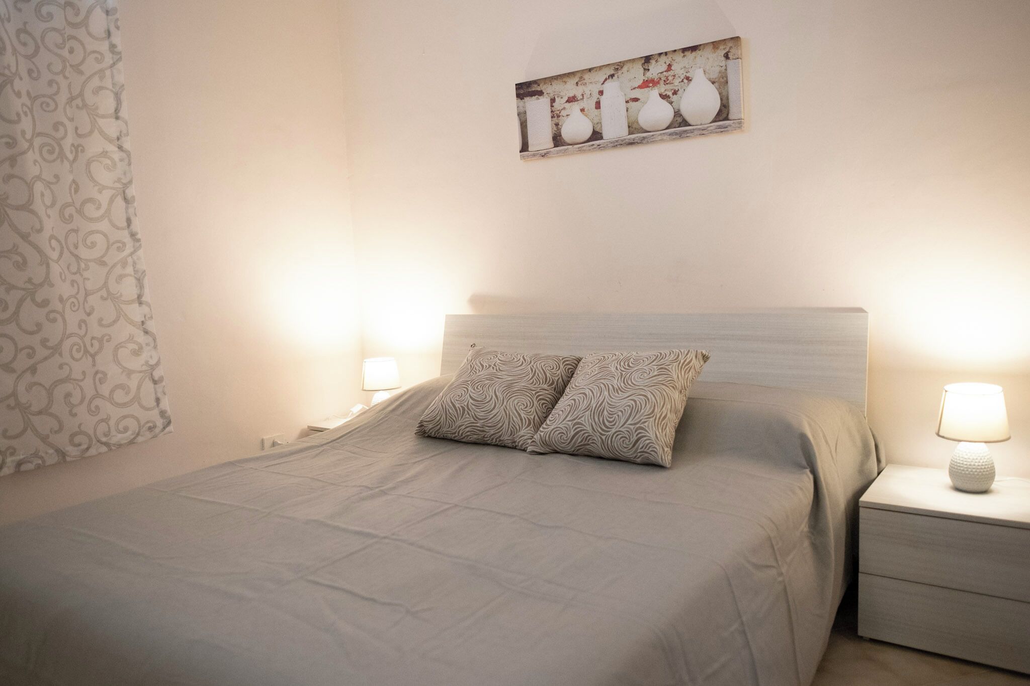 Ein ordentlich bezogenes Bett mit gemusterten Kissen, ein Nachttisch mit Lampe und ein an der Wand hängendes Kunstwerk darüber.