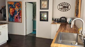 A modern kitchen with dark wood flooring, white cabinets, and a stainless steel sink. There are framed artworks on the walls and a 'Welcome Friends' sign above the sink. - Mystic Breeze Suite 1 (Tarpon Springs)