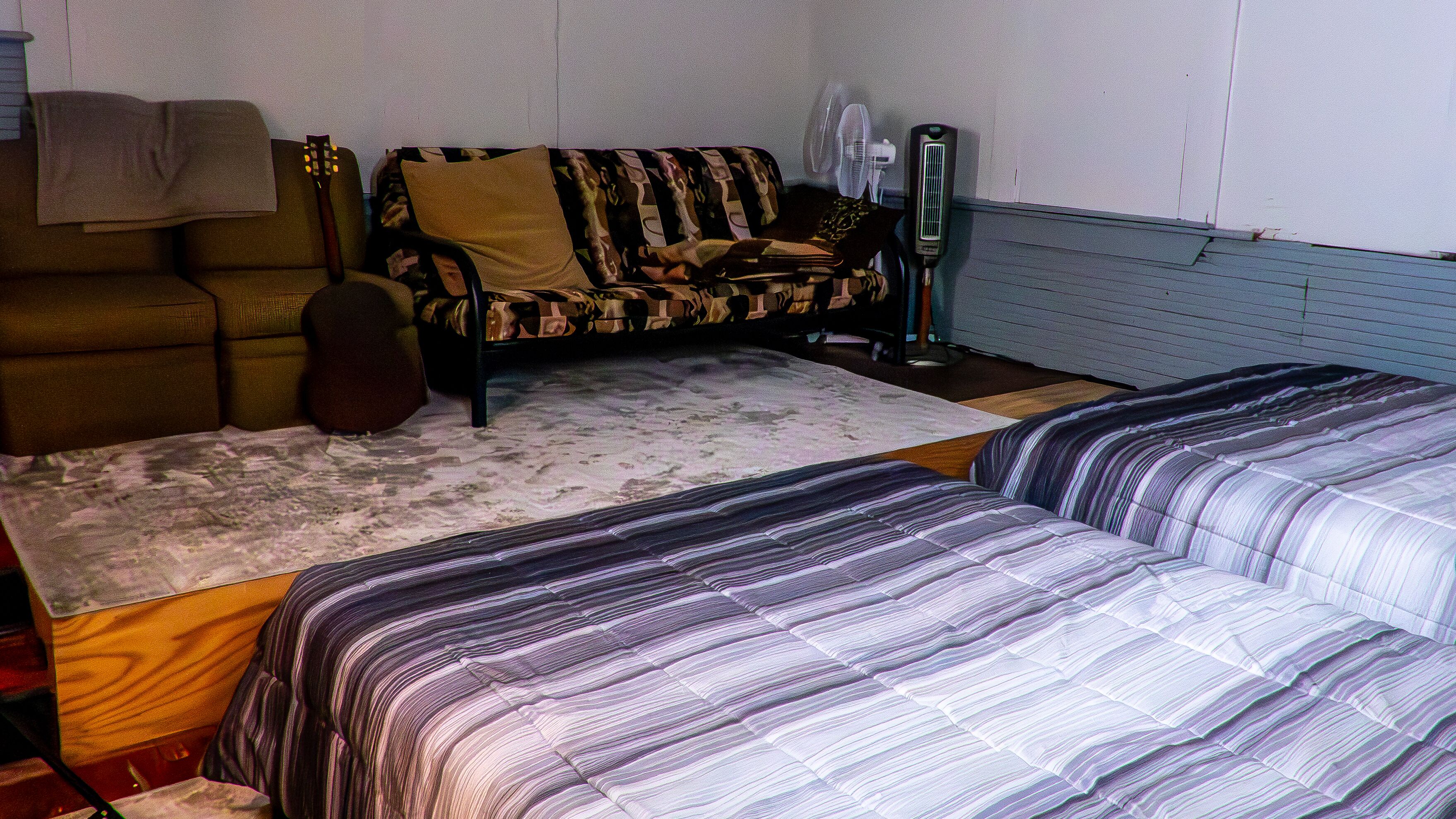 Ein Zimmer mit einer Couch, zwei Betten mit gestreifter Bettwäsche, einem Ventilator und einer Gitarre.