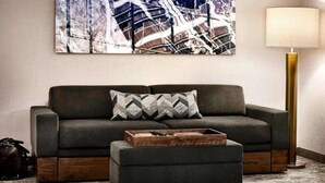 A living room with a grey sofa, a matching ottoman, a patterned throw pillow, a wooden coffee table, a floor lamp, and a large abstract painting on the wall. - Easy Denver Access | Indoor Pool + Free Parking (Parker)