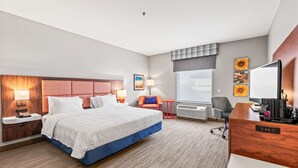 Hotellihuone, jossa on suuri sänky, työpöytä tietokoneella, tuoli, ikkuna, josta on näkymä, ja seinä, jossa on taideteoksia.