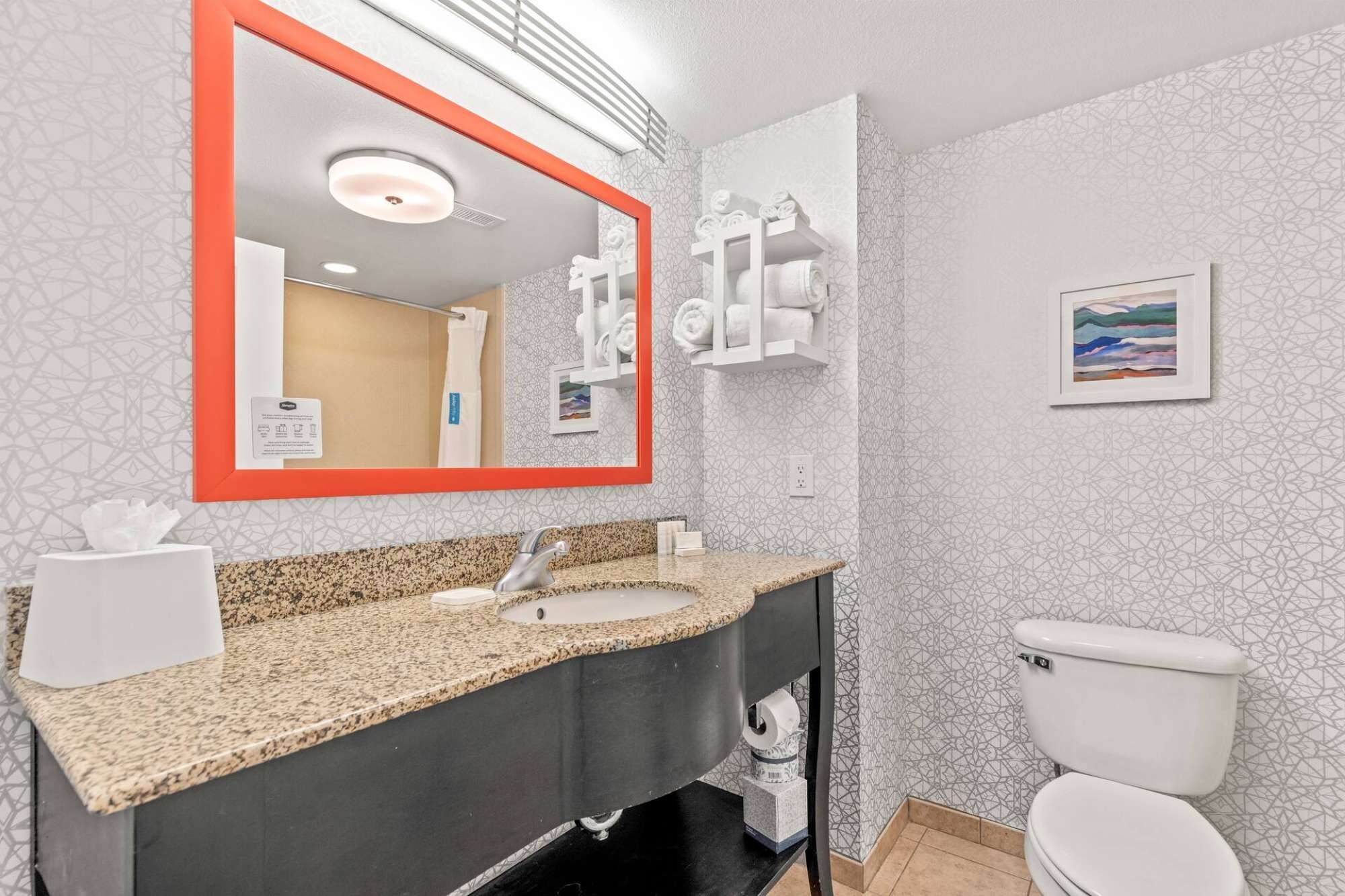 A bathroom with a granite countertop sink, a mirror with a red frame, a toilet, and a towel rack with towels.