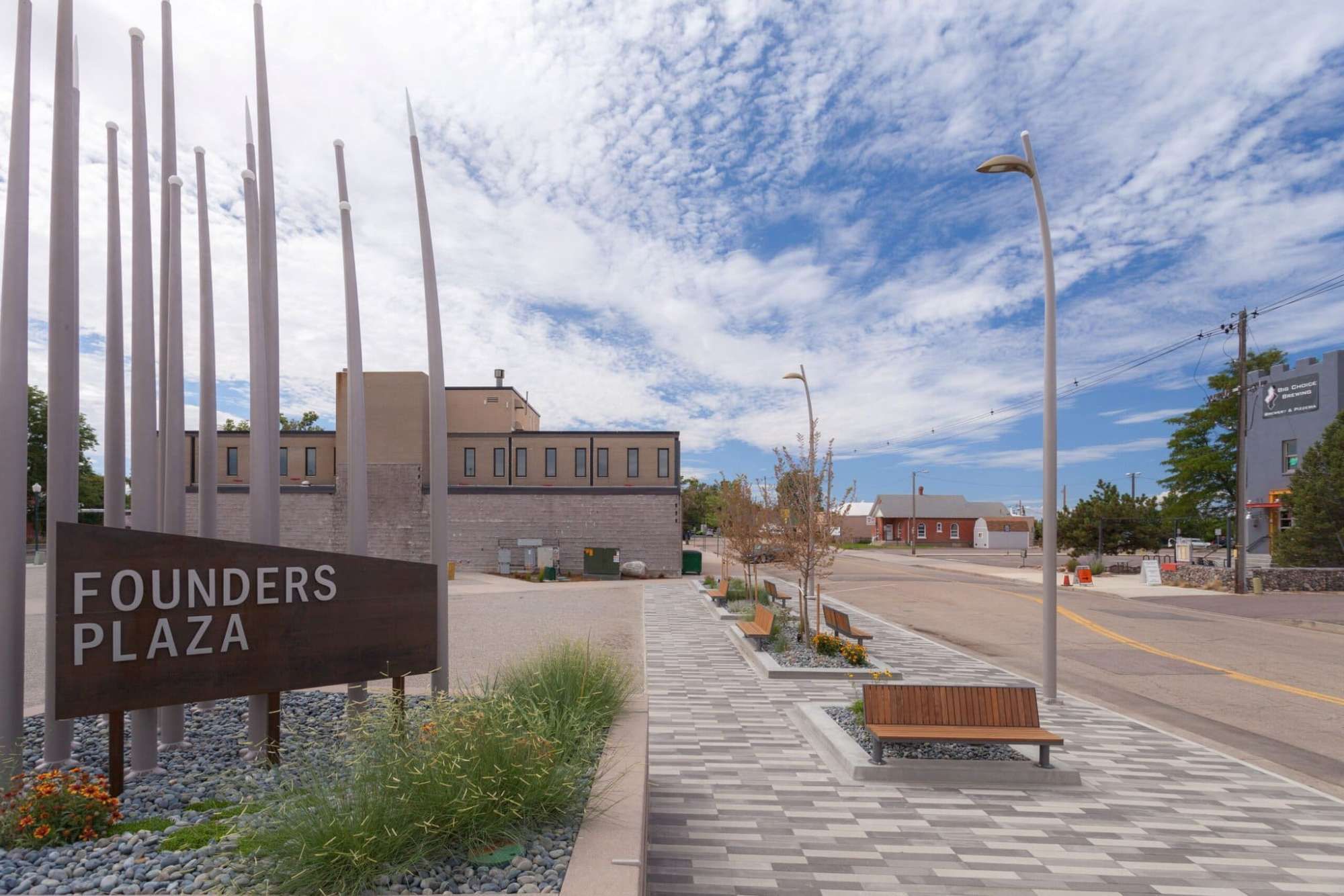 A public plaza with a sign reading 'Founders Plaza,' benches, a patterned walkway, and a building in the background.