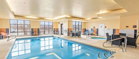 Una piscina interior con agua cristalina, rodeada de sillas de descanso y mesas, y grandes ventanales que ofrecen luz natural.