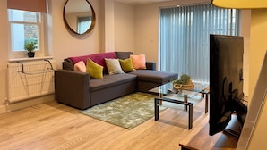 Un salon moderne avec un canapé d’angle, une table basse en verre, un téléviseur à écran plat, un radiateur et une grande fenêtre équipée de stores.