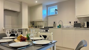 Une cuisine moderne avec des meubles blancs, une table à manger pour quatre personnes et une fenêtre équipée de stores.