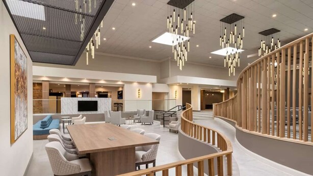 Un amplio vestíbulo de hotel con un gran mostrador de recepción de madera, asientos cómodos, una escalera moderna y luminarias contemporáneas.