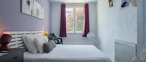 Un dormitorio con cama, mesita de noche, silla, ventana con cortinas y radiador.