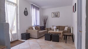 Un salon comprenant un canapé, une table basse et des poufs. Il y a une horloge au mur et une fenêtre avec des rideaux.