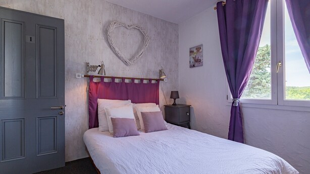 Un dormitorio con cama, una mesita de noche, una lámpara de pared, una decoración con forma de corazón y una ventana con cortinas.