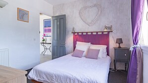 A bedroom with a bed, a nightstand, a lamp, a heart-shaped decoration on the wall, and a door leading to another room. - “Les Nougats” Room with Private Terrace, Shared Garden, and Wi-Fi (Portes-en-Valdaine)