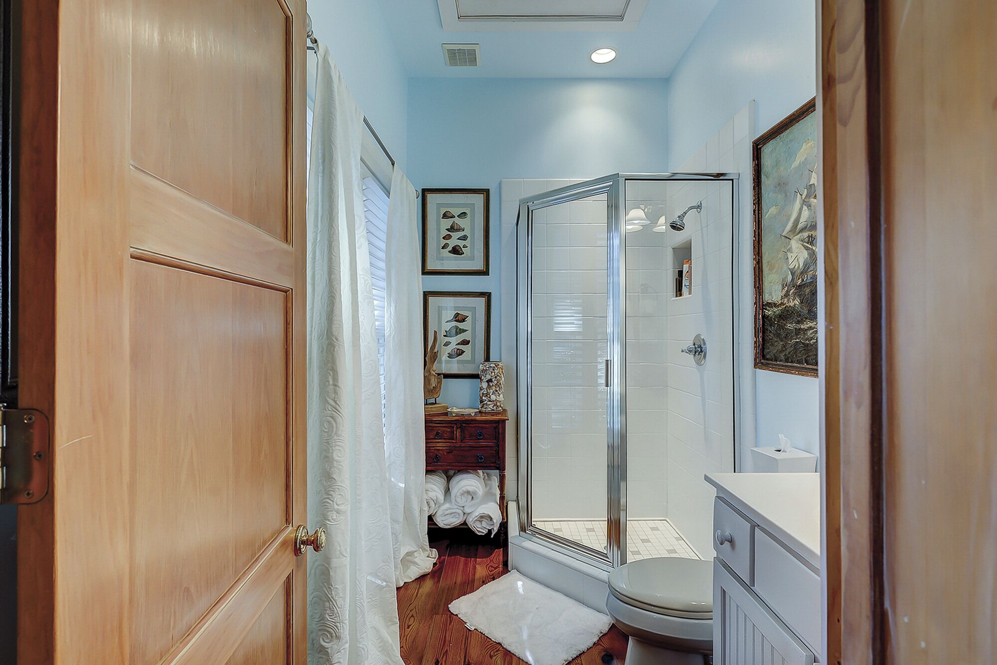 A narrow hallway leading to a bathroom with a glass shower, wooden door, and a small rug on the floor.