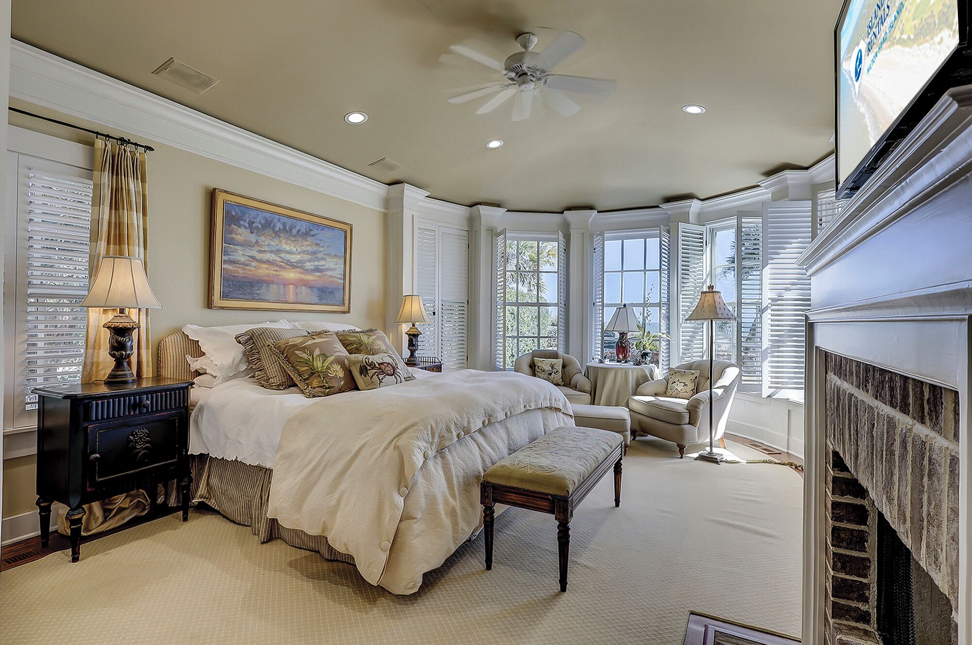 A spacious bedroom with a large bed, a sitting area with a chair and a table, a fireplace, and a ceiling fan.