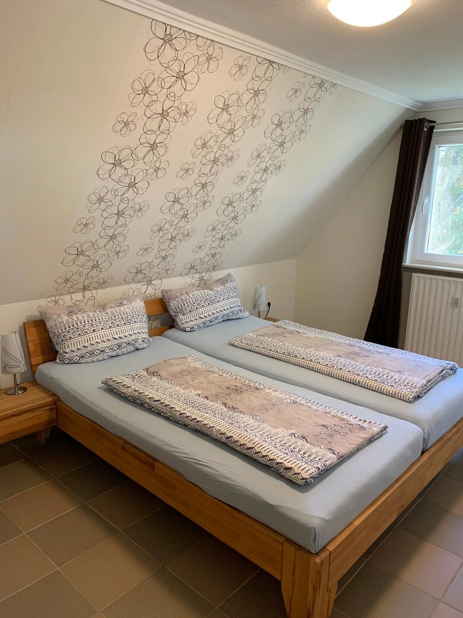 A room with a bed, two pillows, a bedside table, a window, and a wall with a floral pattern.