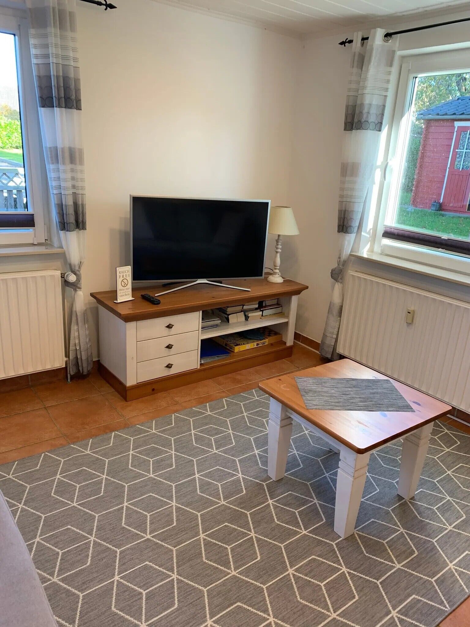A room with a TV on a wooden stand, a small table, a lamp, and a window with curtains.