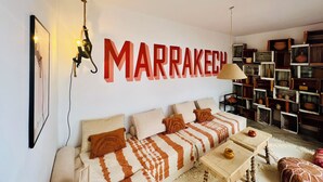 A cozy living room with a large wall-mounted sign reading "MARRAKECH," a sofa with patterned cushions, a wooden coffee table, and a bookshelf filled with various items.
