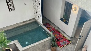 Zona de piscina con mesa y sillas de madera, rodeada de plantas en macetas y un colorido tapete.
