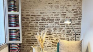Un rincón acogedor con pared de ladrillo, una pequeña mesa redonda con lámpara, una silla con cojín y una estantería con mantas a rayas.