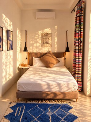 A bedroom with a bed, bedside tables, a wall-mounted air conditioner, a hanging bag, and a patterned curtain. - Private Family-Run Riad • basin • patio (Marrakech)