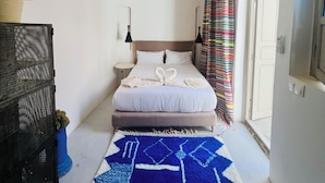 Un dormitorio pequeño y bien iluminado con cama, mesitas de noche, luz de techo, ventana con cortinas y puerta.