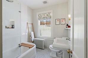 Et badeværelse med badekar, toilet og brusebad. Der er et vindue med udsigt over naturen, en radiator og en hylde med bøger.