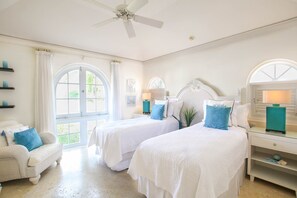 Habitación de hotel con dos camas, un ventilador de techo, una ventana con cortinas y una mesita con lámpara.