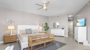 A bedroom with a bed, bedside tables, a bench, a ceiling fan, a dresser, a TV, and a mirror. - Palm Harbor #201 3 Bedroom Condo (Fort Myers Beach)