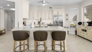 A modern kitchen with white cabinets, a central island with bar stools, a stainless steel refrigerator, and a ceiling fan. - Palm Harbor #201 3 Bedroom Condo (Fort Myers Beach)