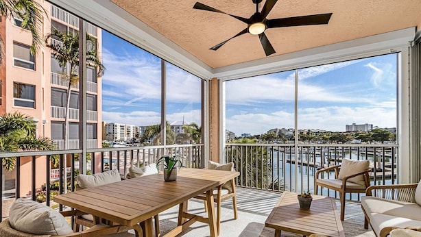 A balcony with a wooden table and chairs, a ceiling fan, and a view of a marina and buildings. - Palm Harbor #201 3 Bedroom Condo (Fort Myers Beach)