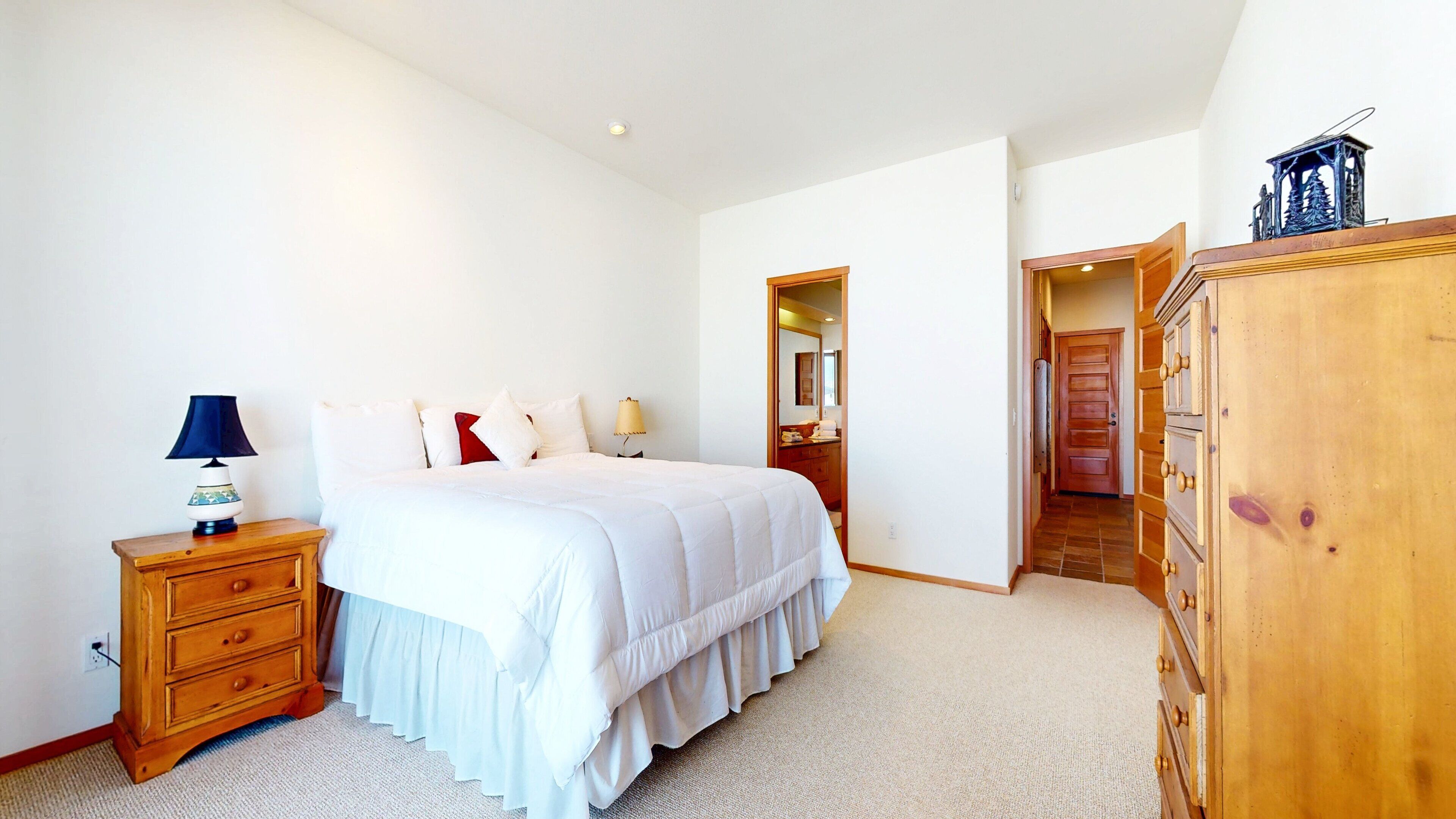 A bedroom with a bed, a dresser, a nightstand, and a lamp. There is a door leading to another room.