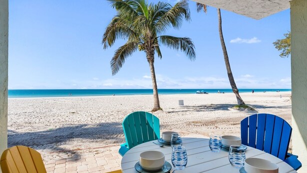 Ein Tisch am Strand, gedeckt mit Tellern, Schüsseln und Gläsern, mit Blick auf einen Sandstrand, Palmen und das Meer.