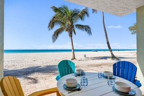 Mesa junto a la playa dispuesta con platos, tazones y vasos, con vista a una playa arenosa con palmeras y el océano.