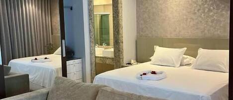 A hotel room with a sofa, two beds, a mirror, and a bathroom visible through a glass door.