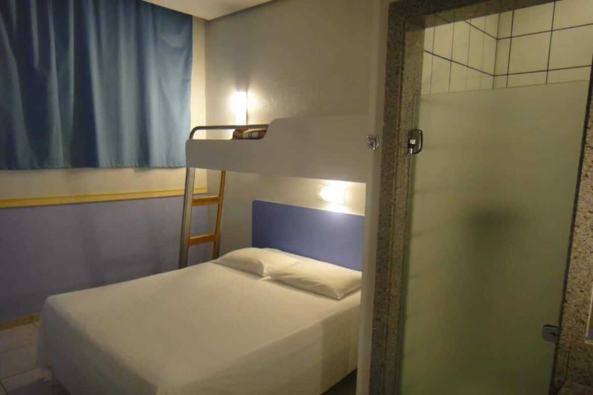 Een enkel bed met hoofdbord, een trap, een raam met blauwe gordijnen en een deur met geëtste glaspanelen.