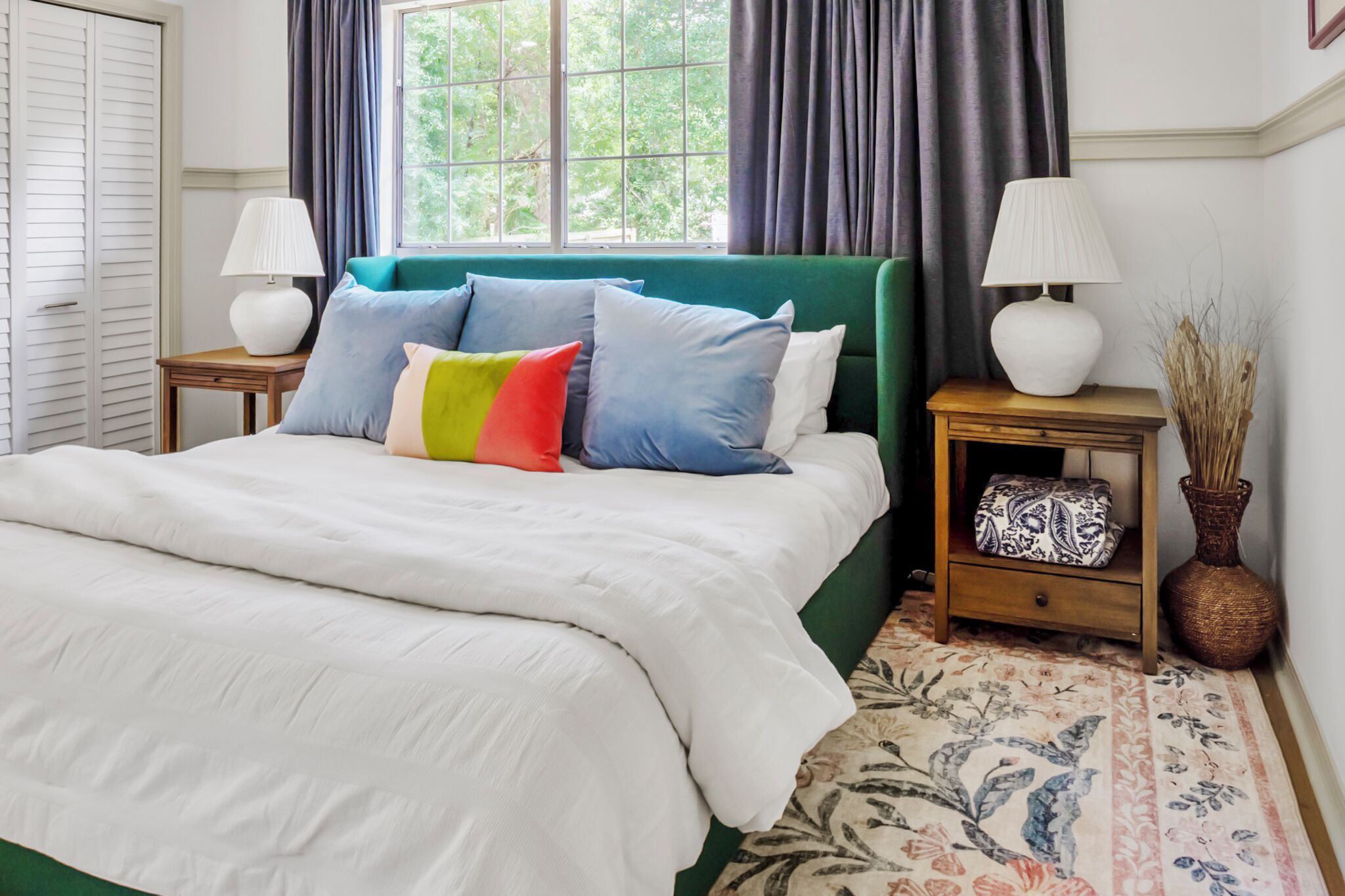 A neatly made bed with a white comforter and multiple pillows, a green upholstered headboard, two bedside tables with lamps, a patterned rug, and a window with curtains.