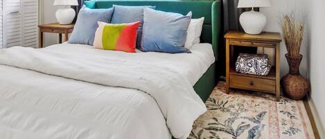 A neatly made bed with a white comforter and multiple pillows, a green upholstered headboard, two bedside tables with lamps, a patterned rug, and a window with curtains.