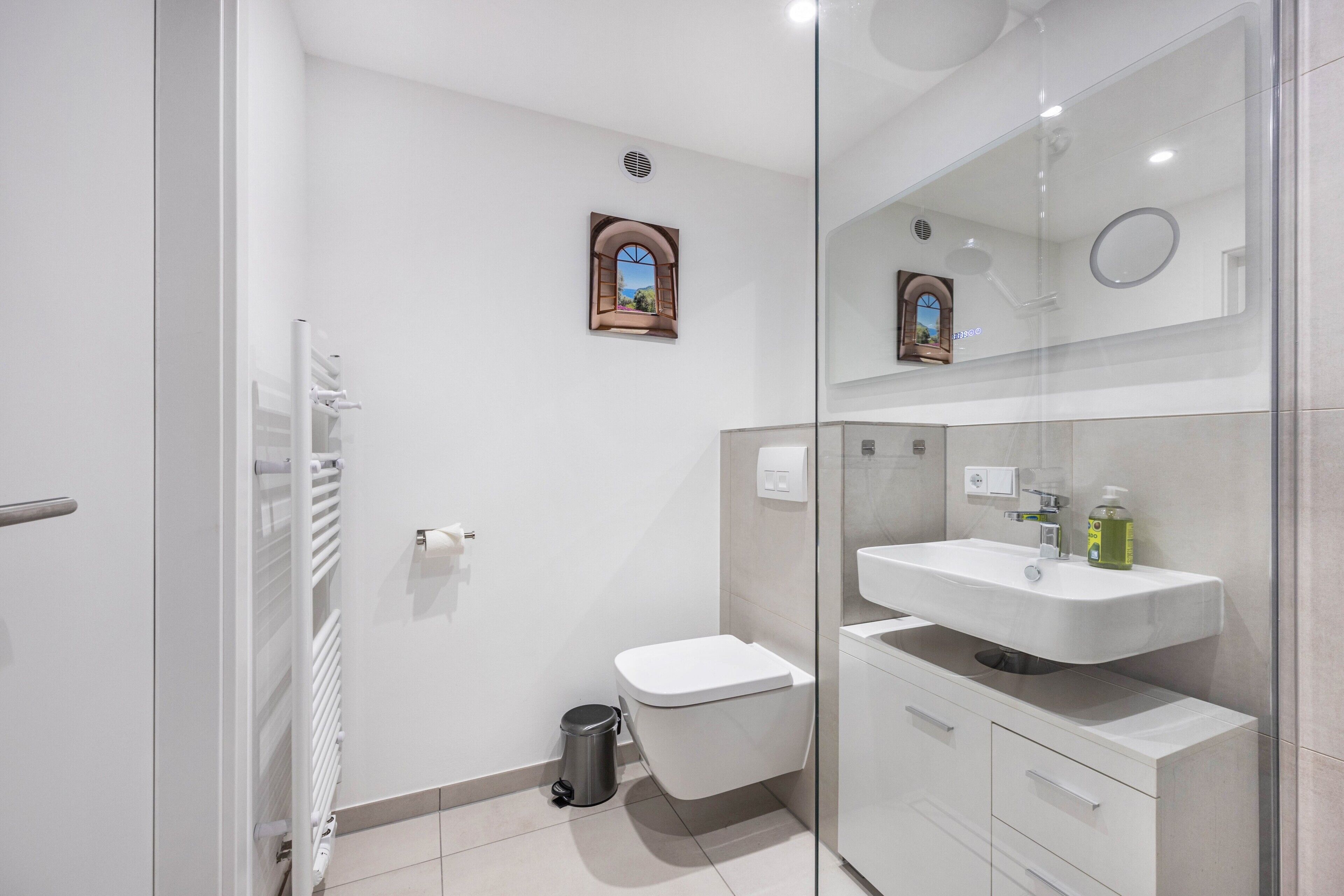 Ein modernes Badezimmer mit Duschkabine aus Glas, Toilette in Weiß, Waschtisch und einem kleinen Fenster mit Blick.