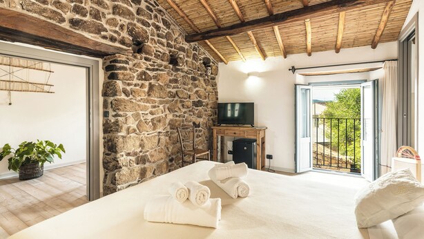 Un dormitorio con paredes de piedra, techo de madera, cama con ropa blanca, televisor y balcón con vistas.