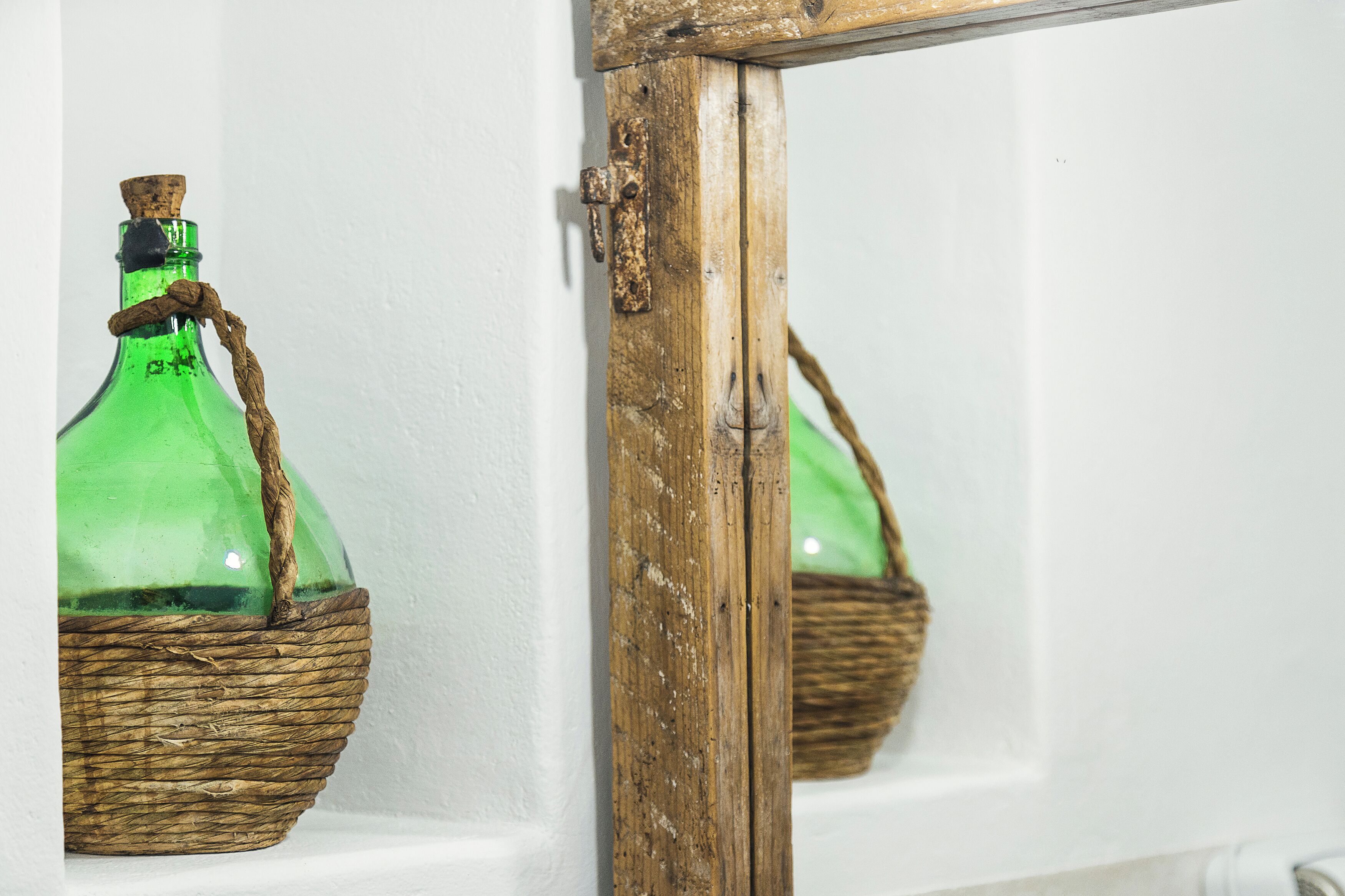 Una botella de vidrio verde con asa de cuerda, colocada en una cesta tejida frente a una pared blanca.