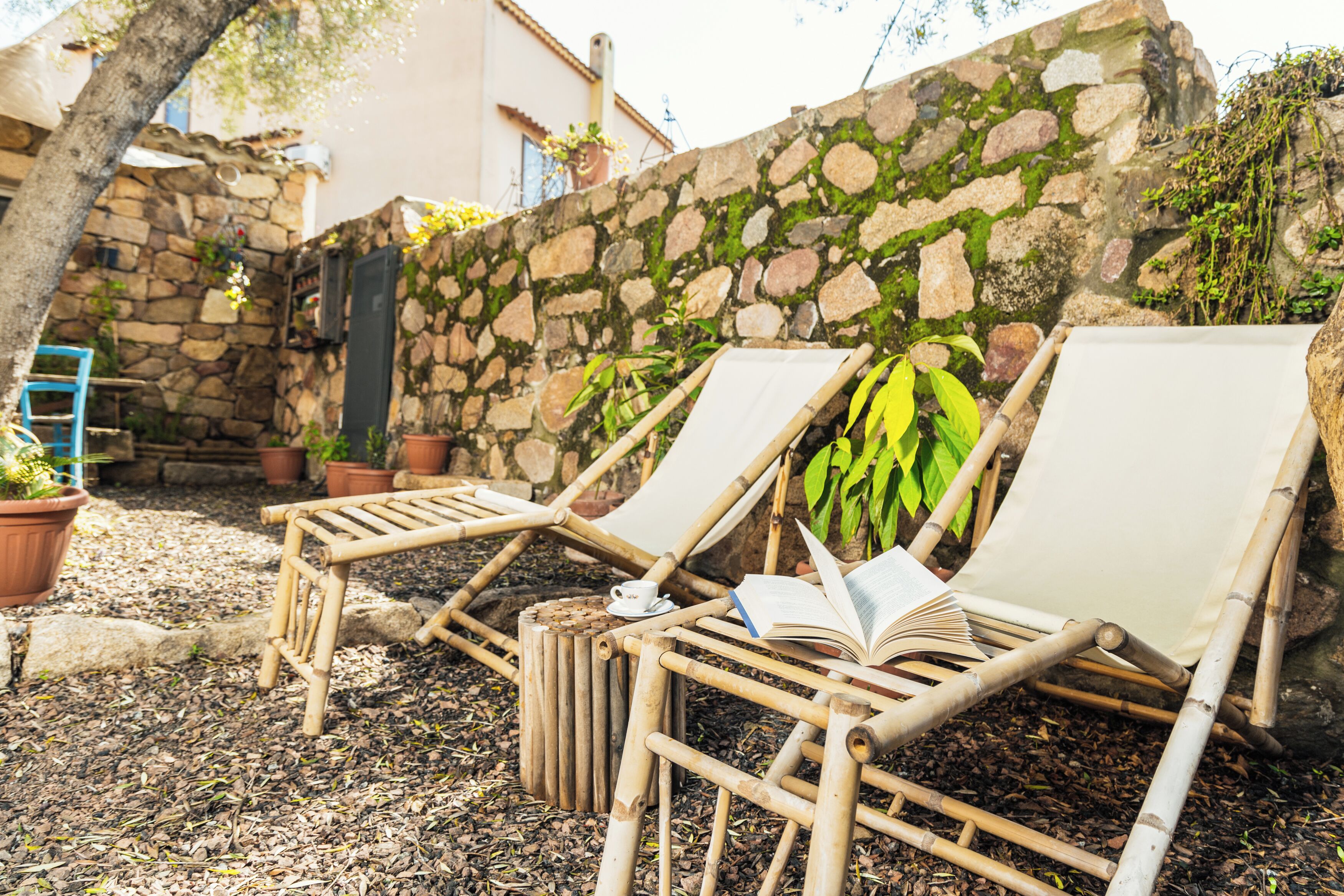 Two bamboo lounge chairs with a book and a cup, set against a stone wall with potted plants.