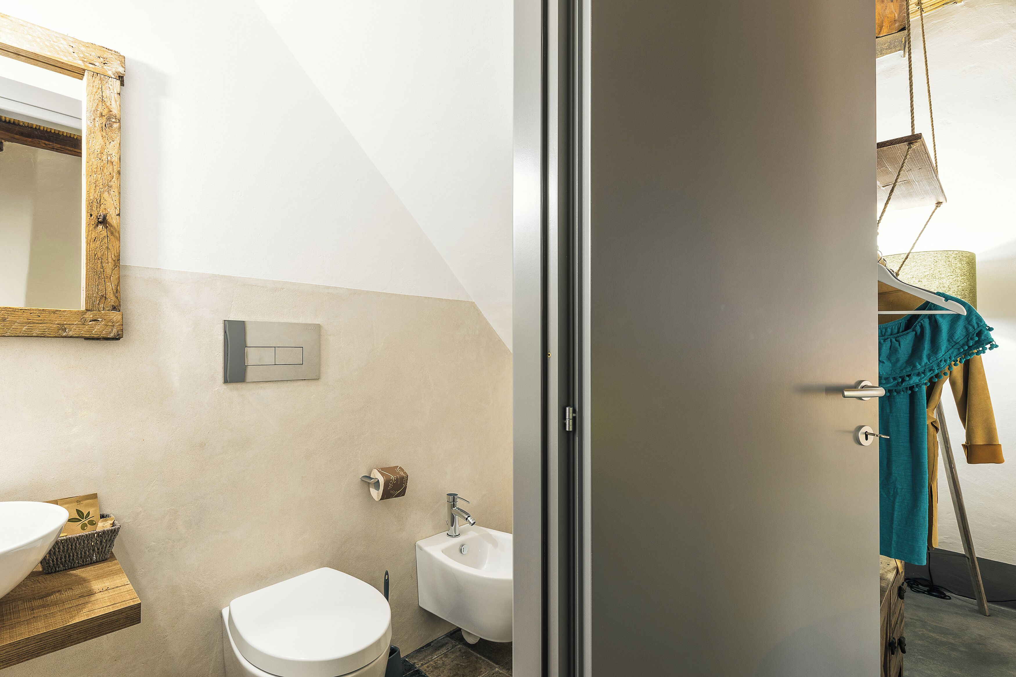 A bathroom with a white sink, a toilet, and a bidet. There is a wooden shelf with a bowl and a towel hanging on the wall.