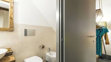 A bathroom with a white sink, a toilet, and a bidet. There is a wooden shelf with a bowl and a towel hanging on the wall.