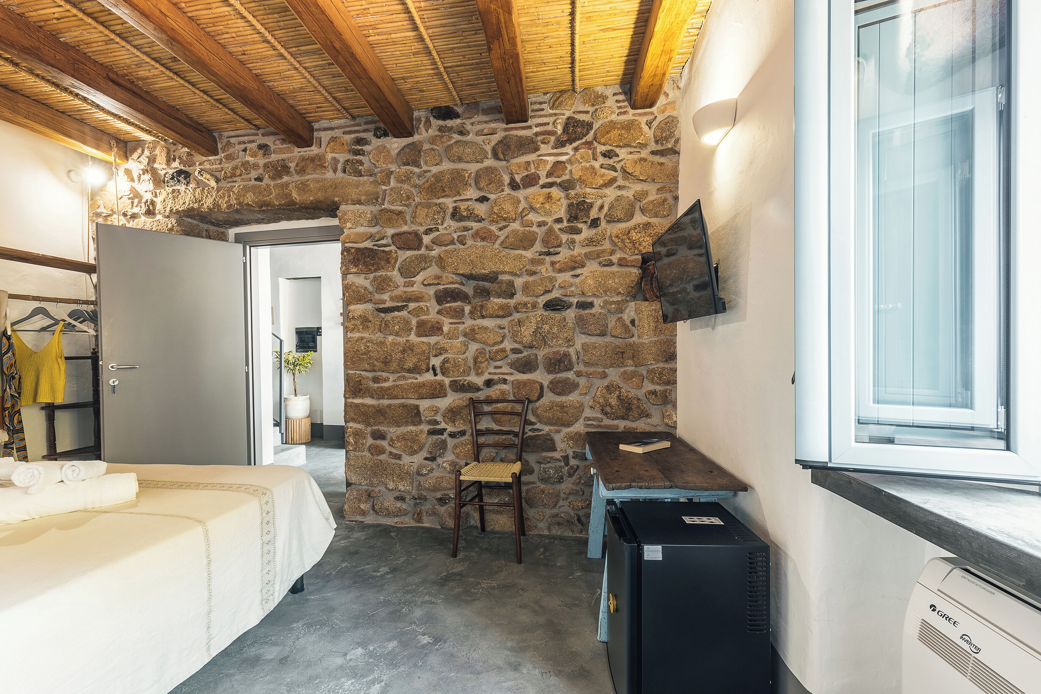 Habitación con paredes de piedra, una cama, una silla, una mesita y un ventanal.