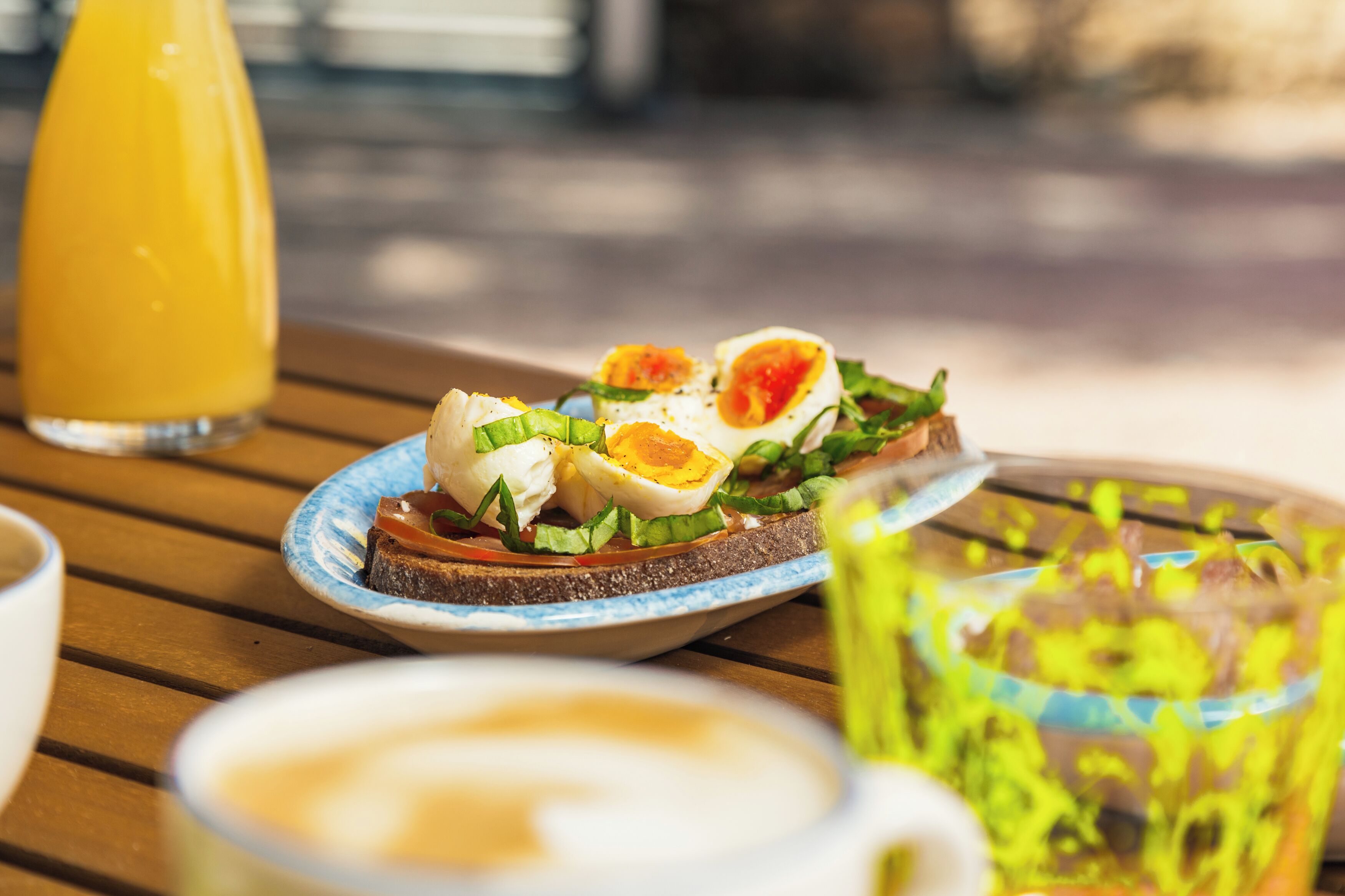 Desayuno con tostadas cubiertas de huevos y vegetales verdes, un vaso de jugo de naranja y una taza de café sobre una mesa de madera.