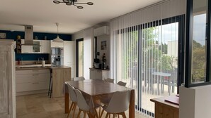 Una cocina moderna con armarios blancos, una zona de comedor con mesa y sillas de madera, y amplias ventanas con persianas.