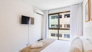 Ένα δωμάτιο ξενοδοχείου με ένα κρεβάτι, μια τηλεόραση επίπεδης οθόνης τοποθετημένη στον τοίχο και ένα μεγάλο παράθυρο με θέα σε ένα κτίριο.