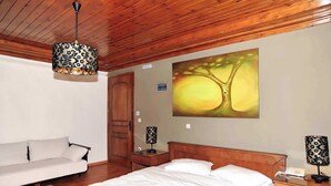 A bedroom with a wooden ceiling, a bed with white linens, a wooden nightstand, a lamp, a painting of a tree, and a white sofa. - Goura Guesthouse Mountain Double Suite Lake Doxa (Goura)