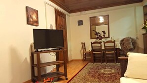 A living room with a flat-screen TV on a wooden stand, a dining table with chairs, a mirror, and a patterned rug. - Goura Guesthouse Family Suite Lake Doxa (Goura)