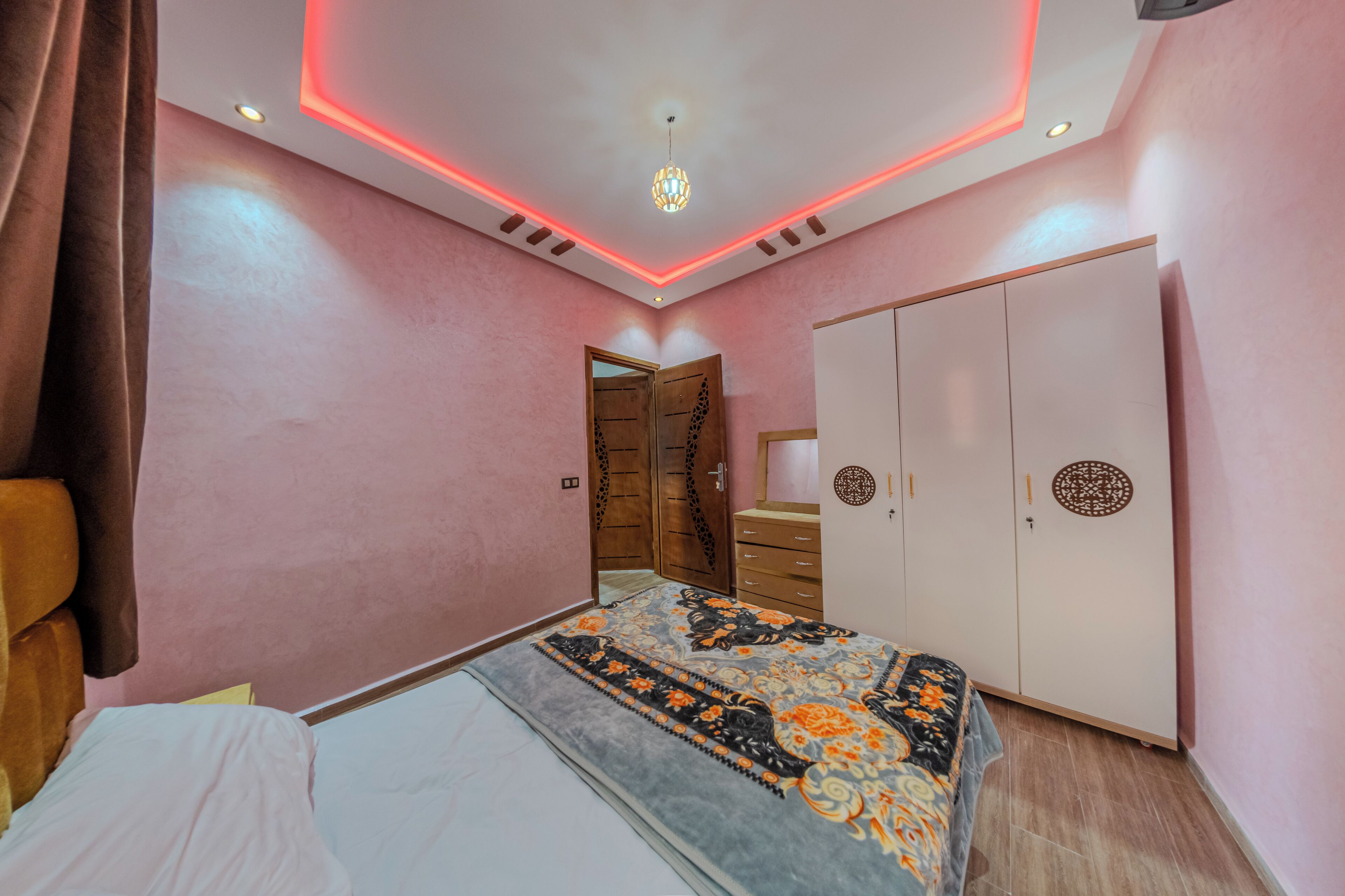 Quarto com paredes cor-de-rosa, cama com colcha estampada, guarda-roupa e iluminação vermelha no teto.